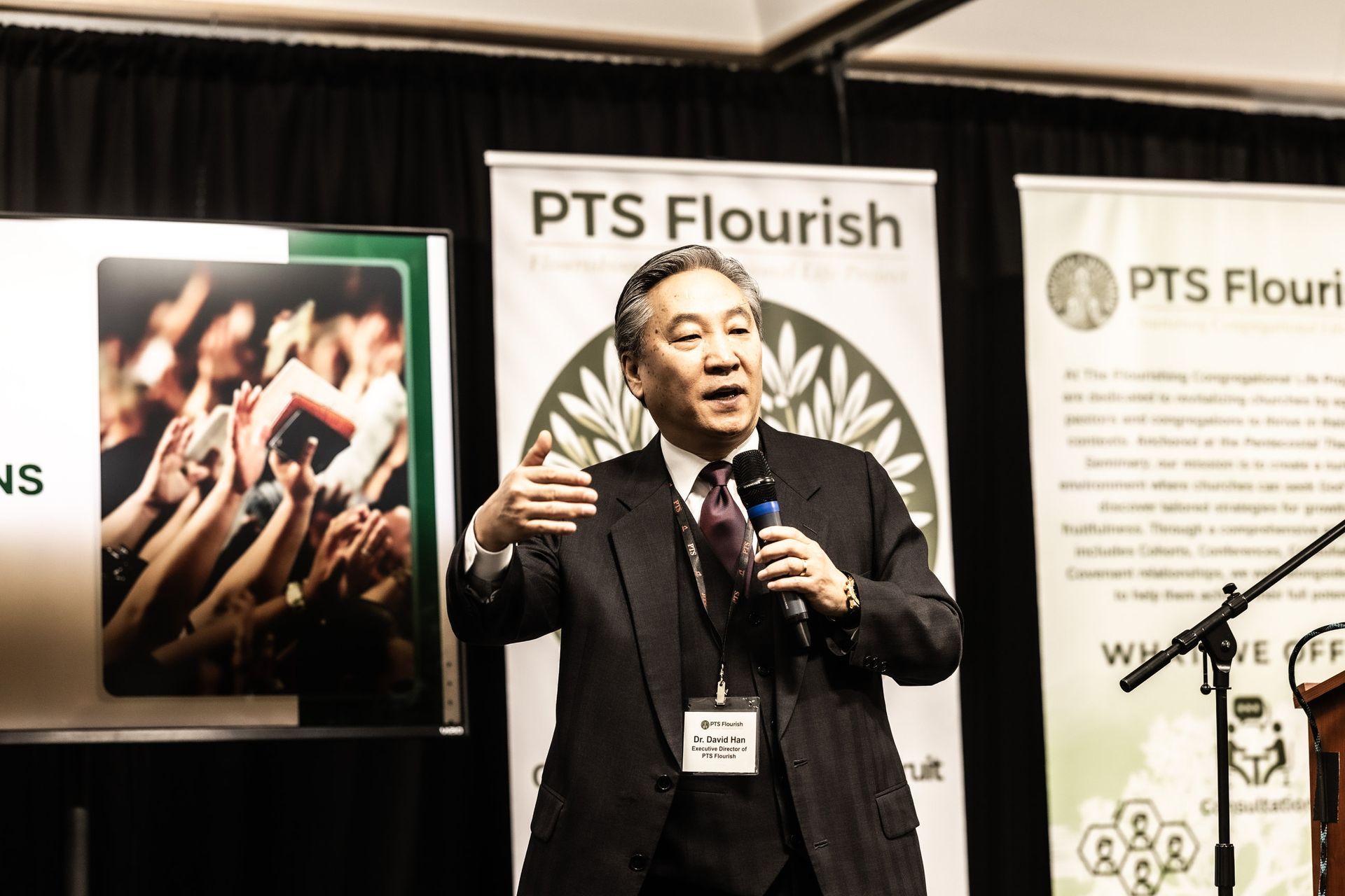 A man in a suit and tie is speaking into a microphone in front of a sign that says pts flourish