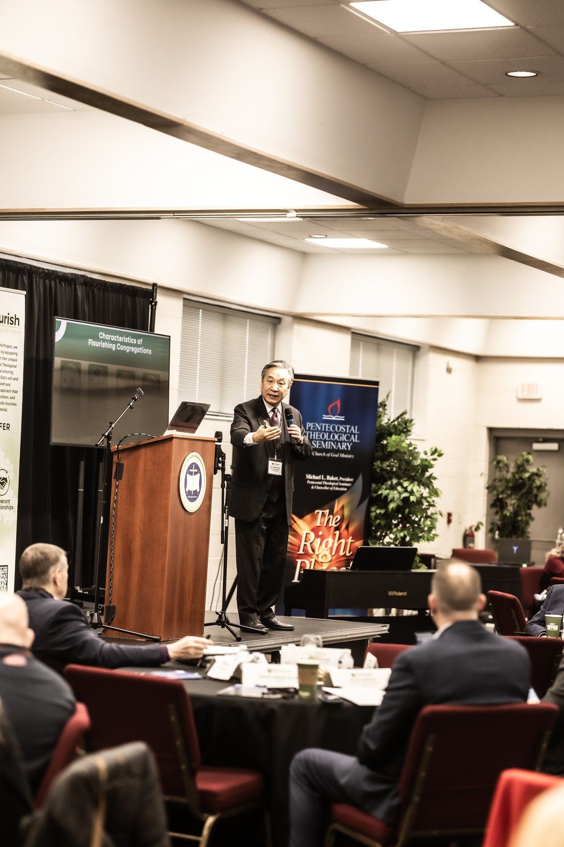 A man is standing at a podium giving a presentation to a group of people sitting at tables.