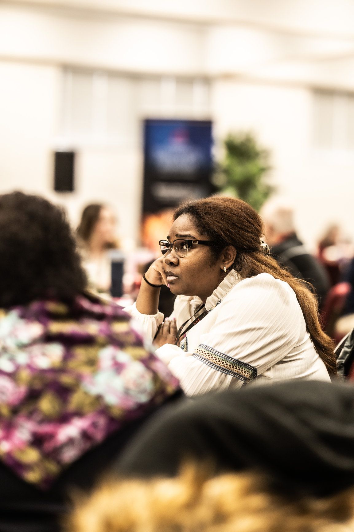 A woman wearing glasses is sitting in a room with other people.