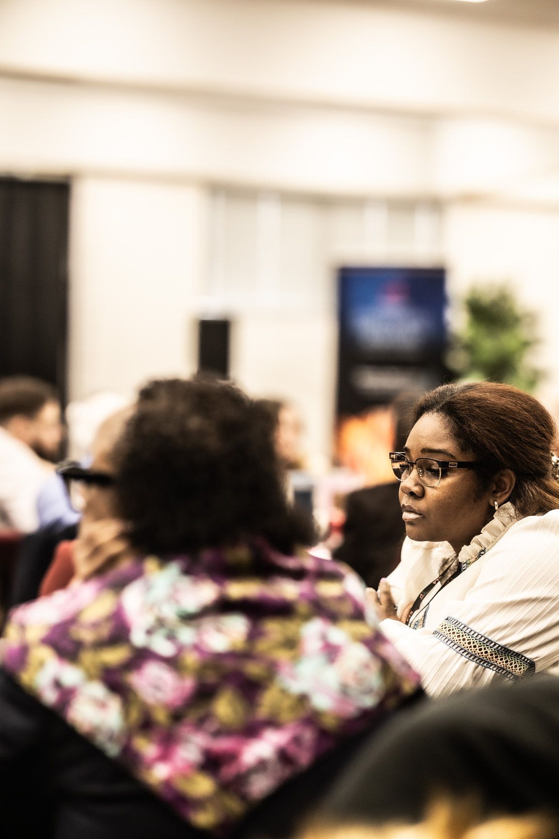 A woman wearing glasses is sitting in a room with other people.