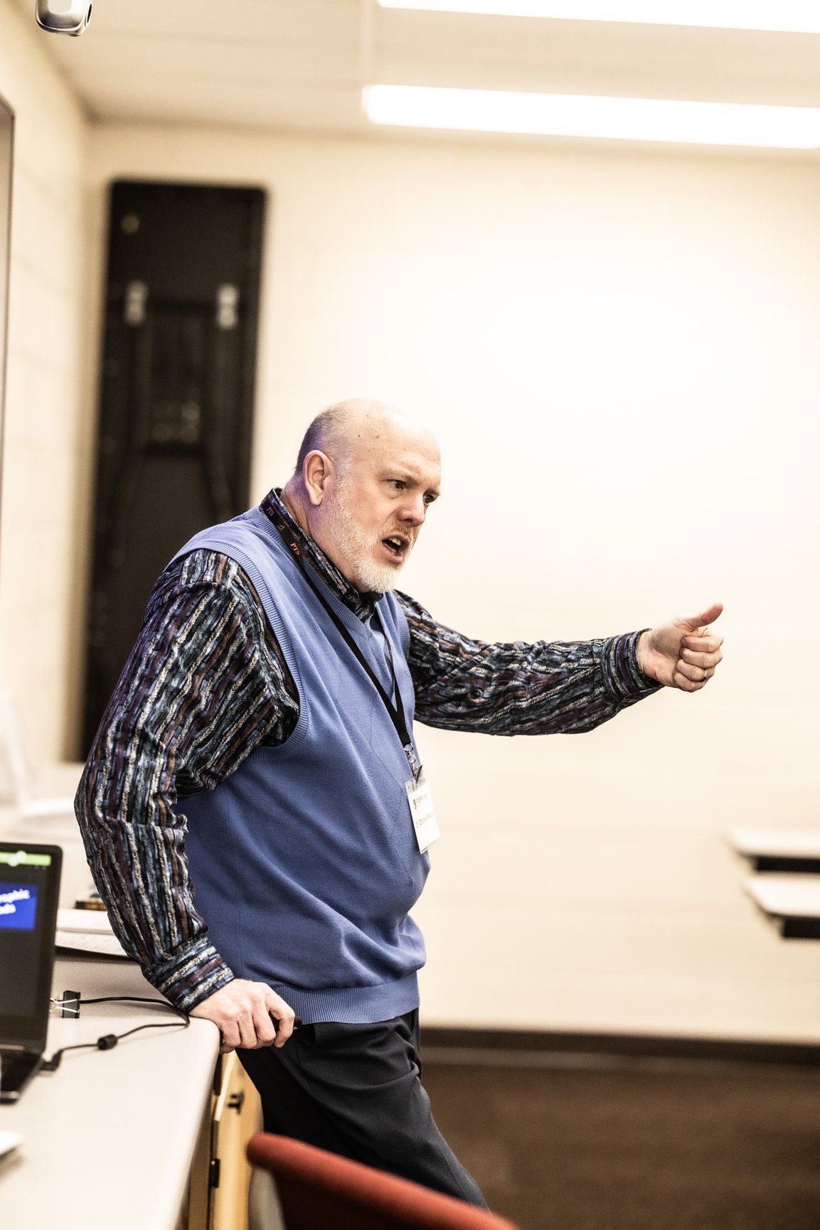 A man in a blue vest is giving a presentation in a classroom.