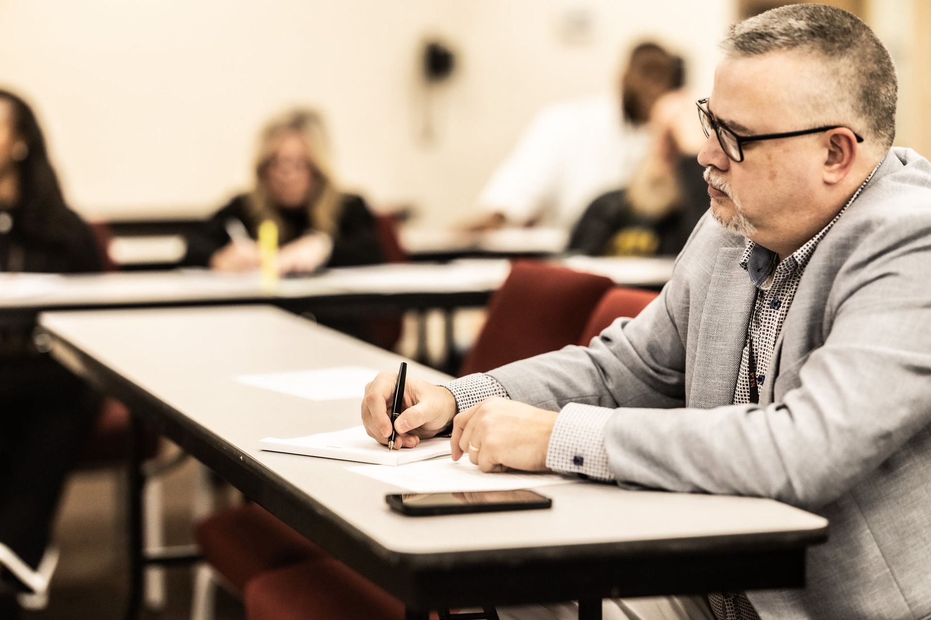A man is sitting at a table in a classroom writing on a piece of paper.