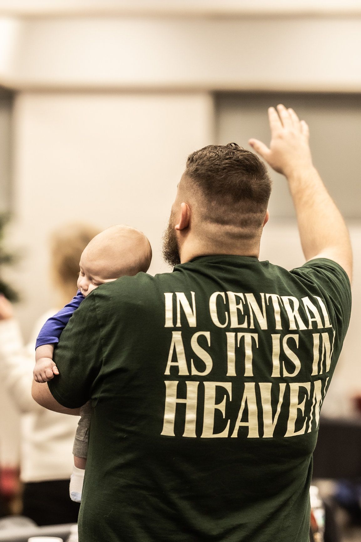 A man holding a baby wearing a shirt that says in central as it is in heaven