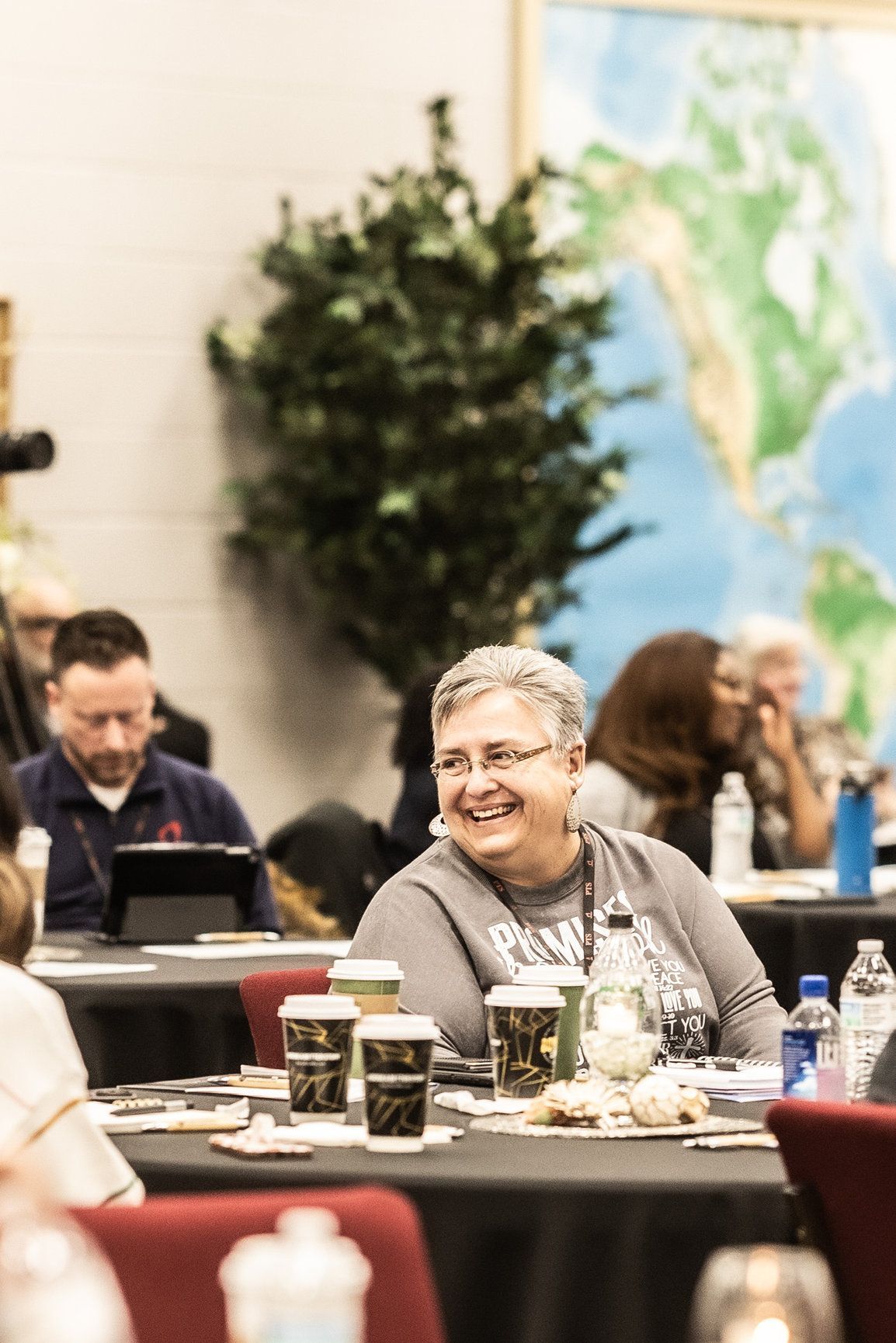 A group of people are sitting at tables in a room with a map of the world in the background.