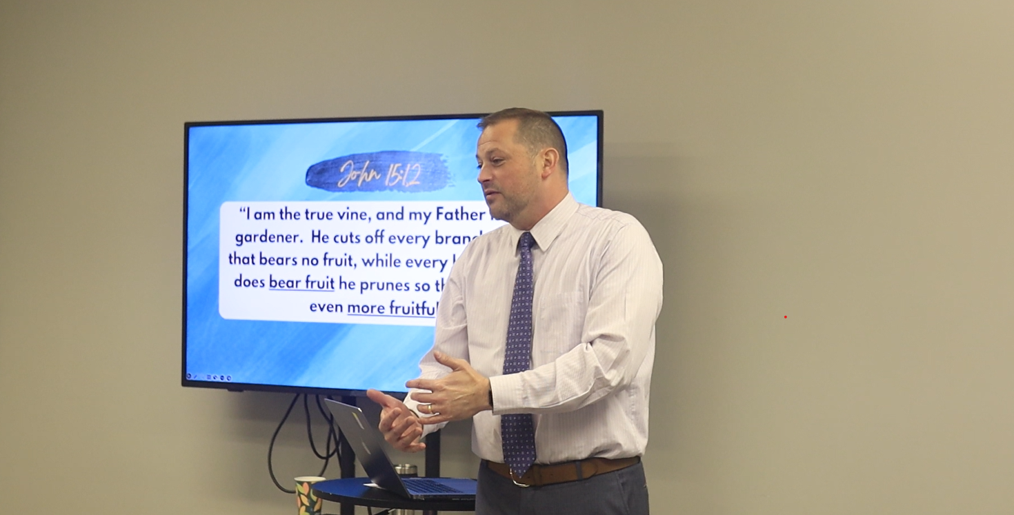A man in a white shirt and tie is giving a presentation in front of a large screen.