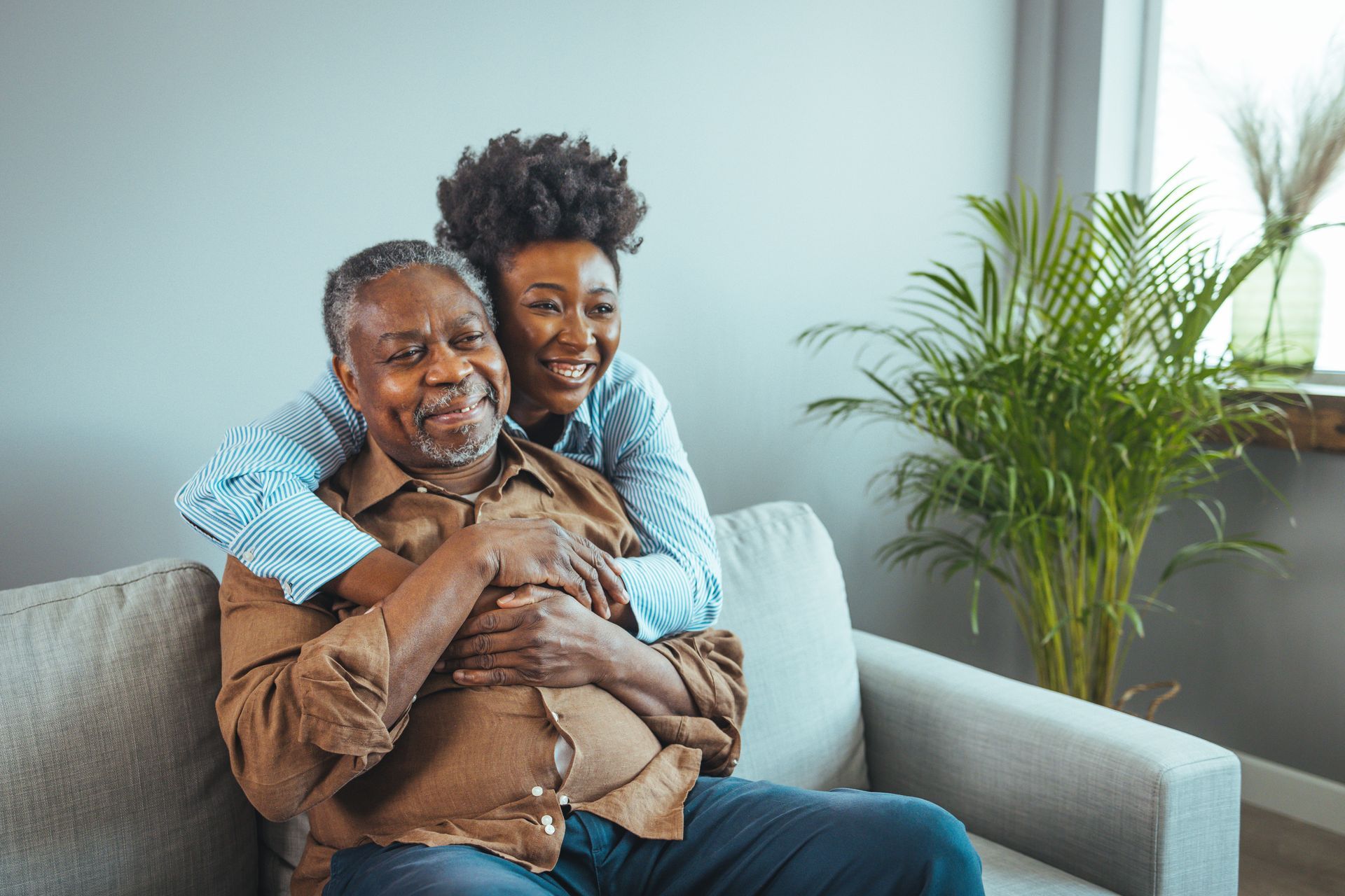 adult daughter smiles and hugs her father from behind while hes sitting on couch