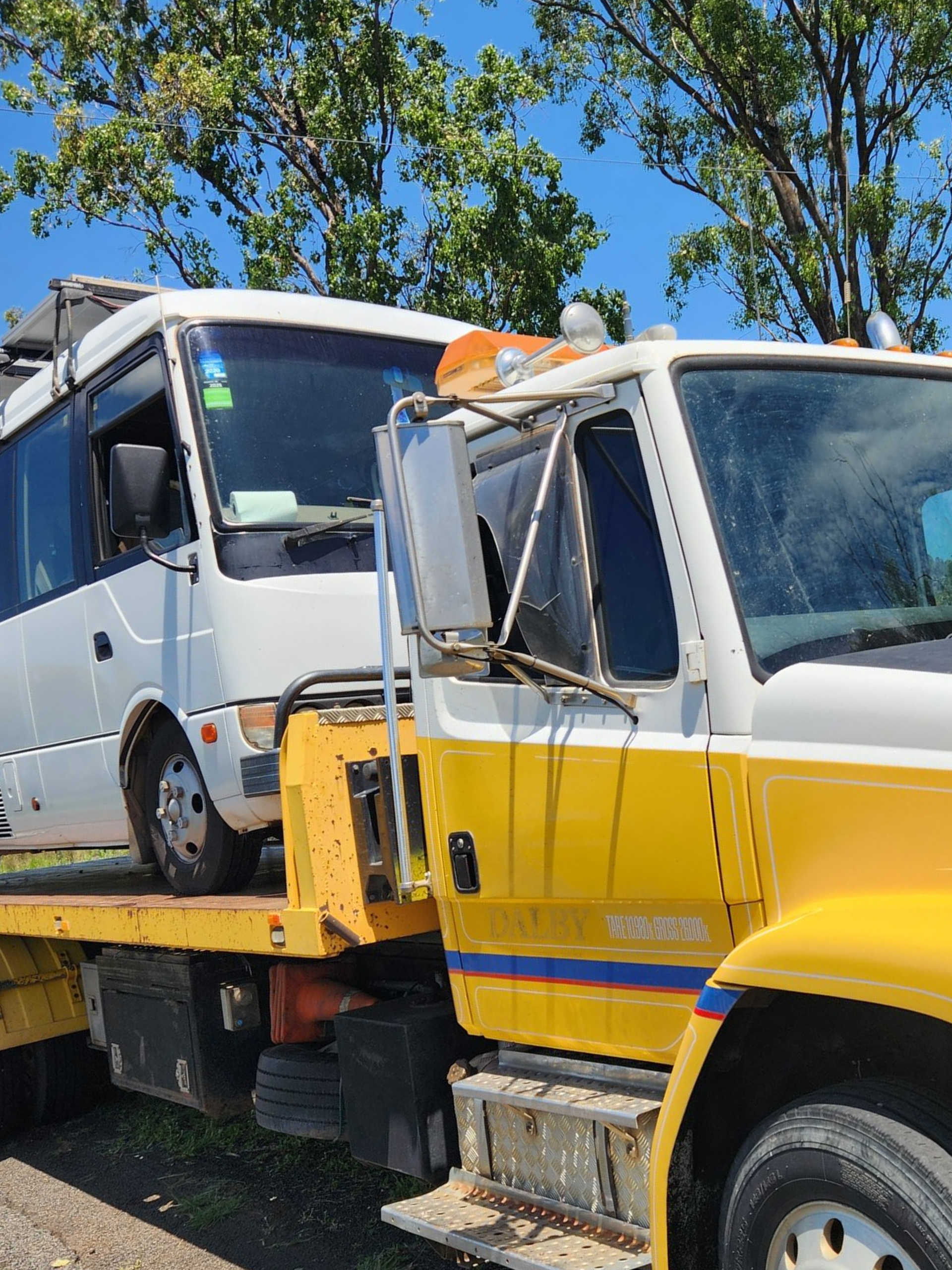 White Bus Loaded On A Yellow Tow Truck, Parked Outdoors With Trees In The Background — Downs Towing & Recovery Service in Moonie, QLD