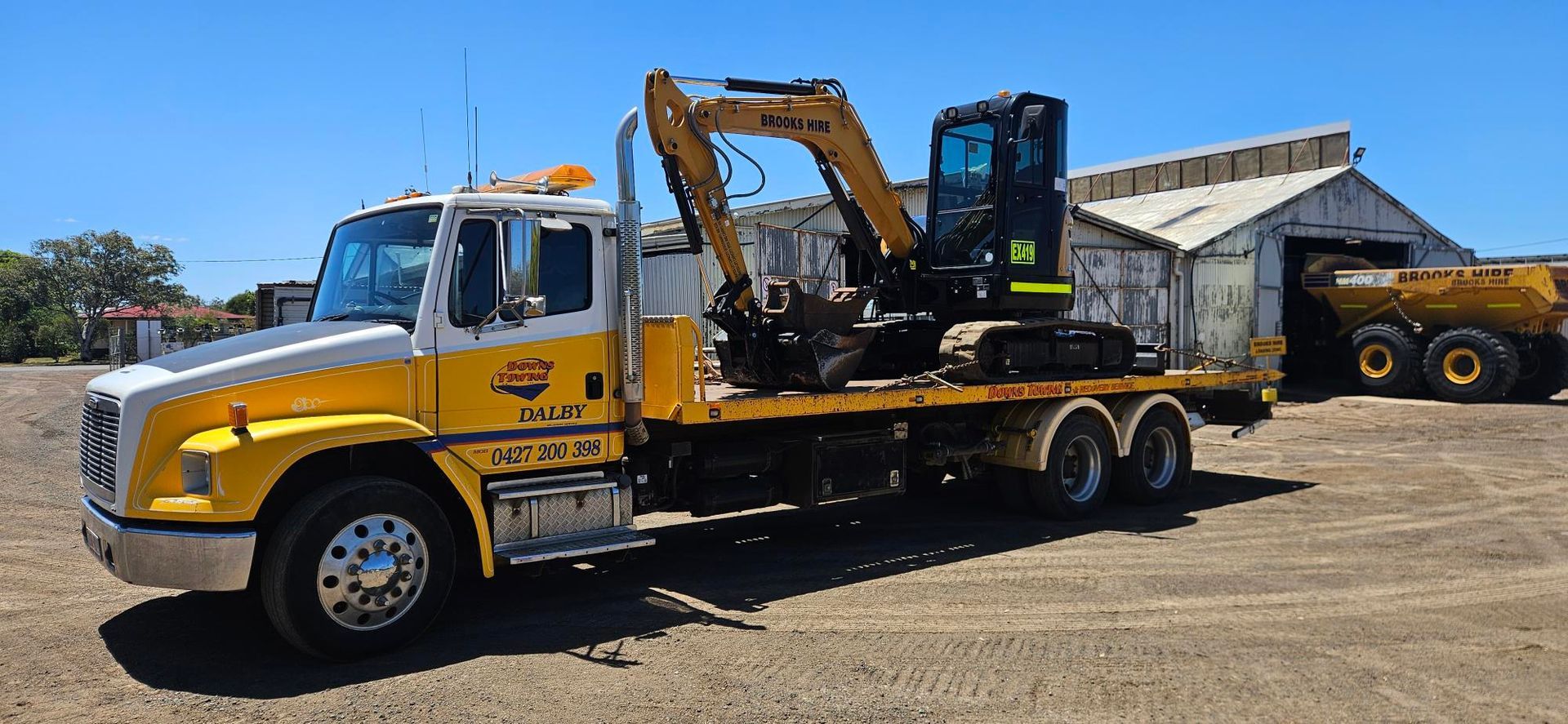 Yellow Tow Truck Hauling A Compact Excavator In A Gravel Lot Under A Blue Sky — Downs Towing & Recovery Service in Miles, QLD