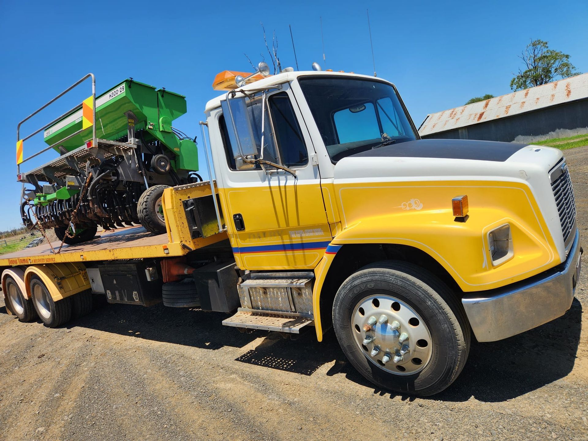 Yellow And White Flatbed Truck Carrying Agricultural Equipment On A Dirt Road — Downs Towing & Recovery Service in Dalby, QLD
