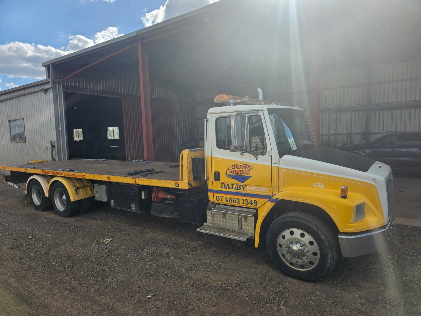 Yellow Flatbed Tow Truck Parked Beside A Warehouse Under Bright Sunlight — Downs Towing & Recovery Service in St. George, QLD