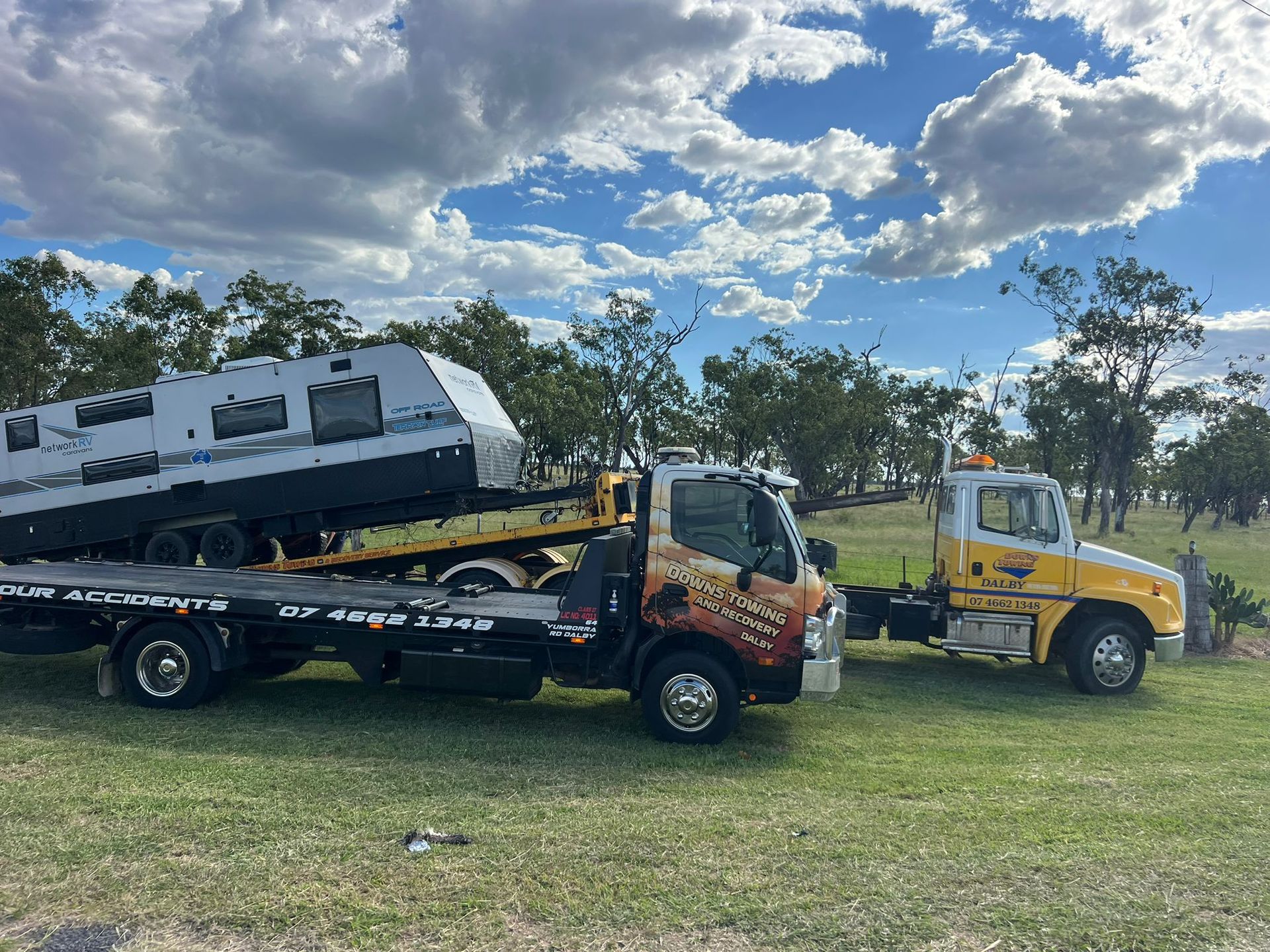 Tow Trucks Hauling A White Camper And A Yellow Truck On A Grassy Field Under A Cloudy Sky — Downs Towing & Recovery Service in Moonie, QLD
