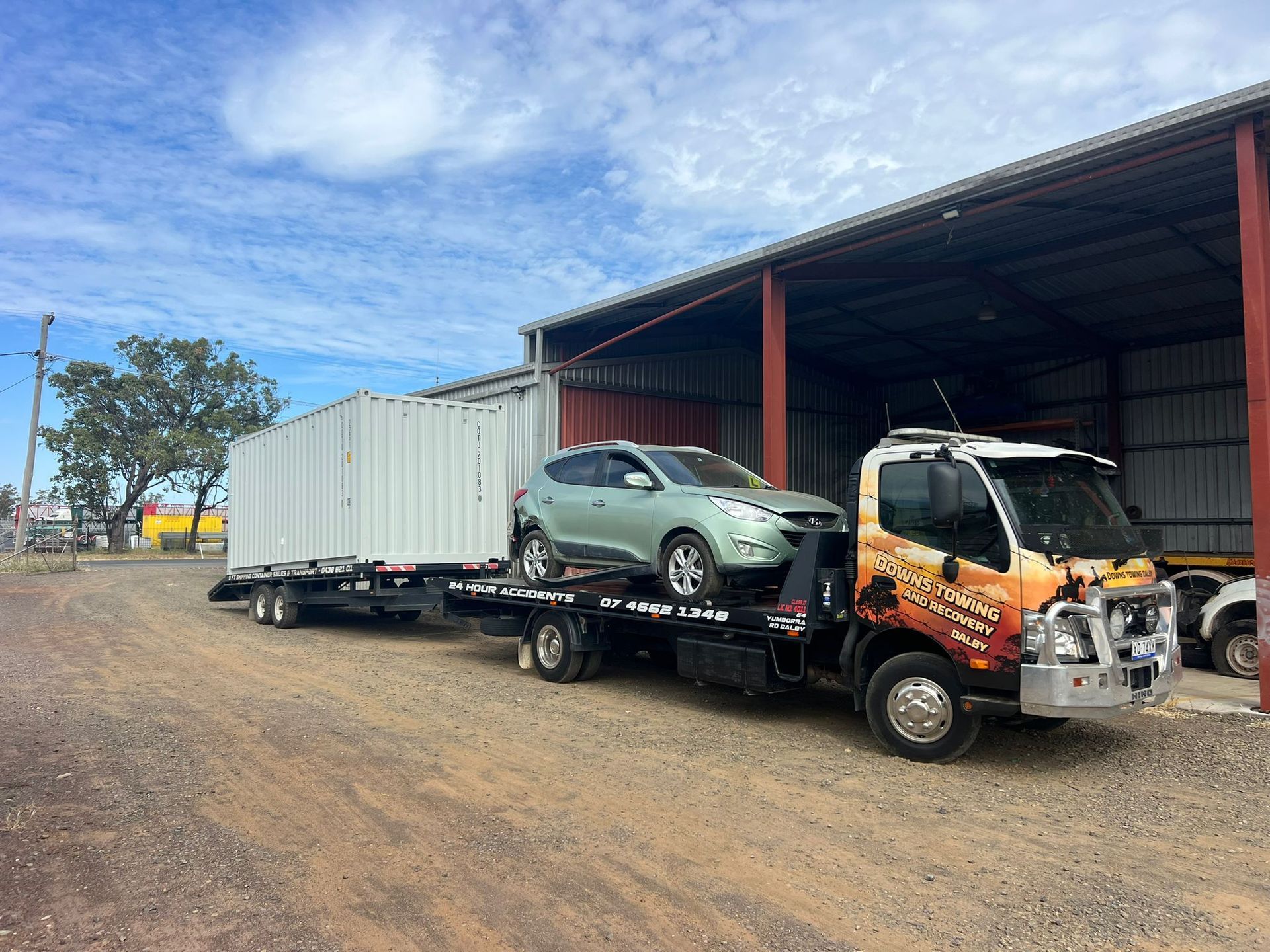Tow Truck Hauling A Green Car And White Shipping Container Beside A Metal Shed On A Dirt Lot — Downs Towing & Recovery Service in Moonie, QLD