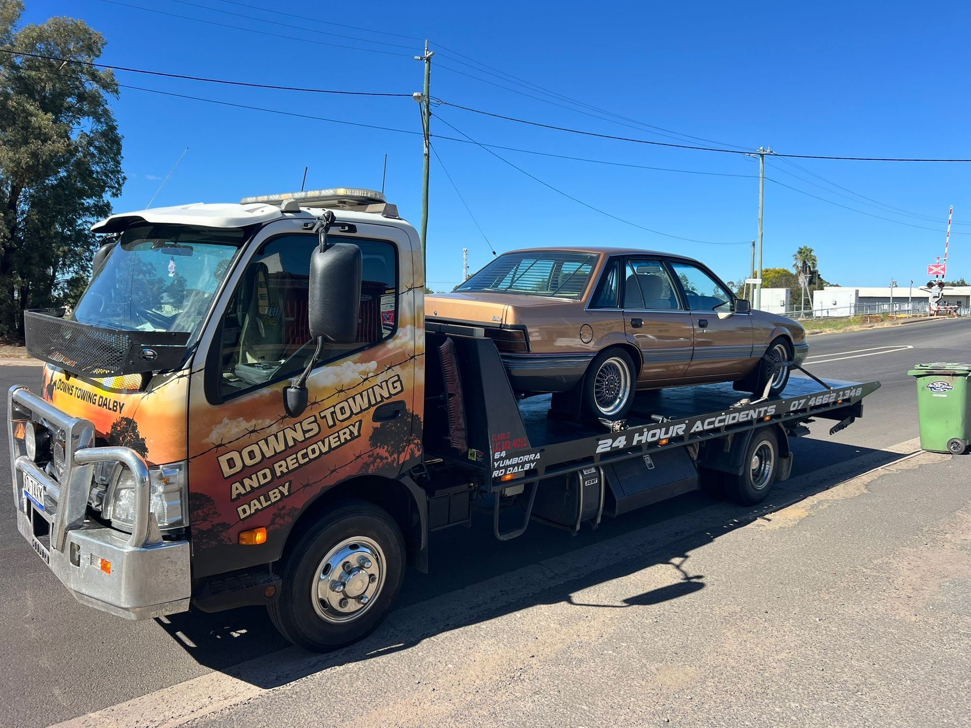 Tow Truck Hauling A Brown Sedan On A Flatbed Along A Roadside Under A Clear Blue Sky — Downs Towing & Recovery Service in Dalby, QLD