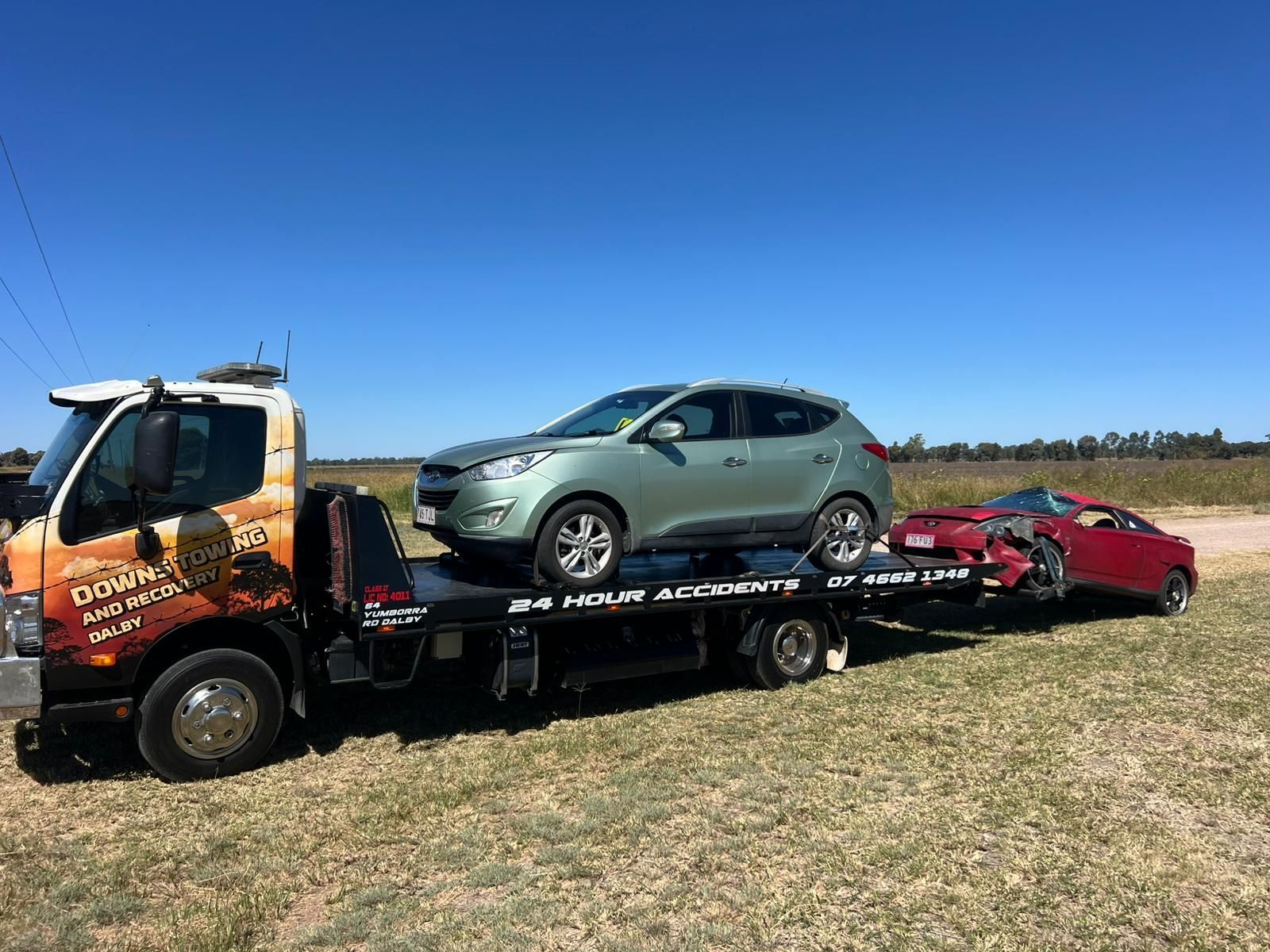 Tow Truck Carrying A Green SUV And Red Sports Car Across A Grassy Field Under A Blue Sky — Downs Towing & Recovery Service in Dalby, QLD