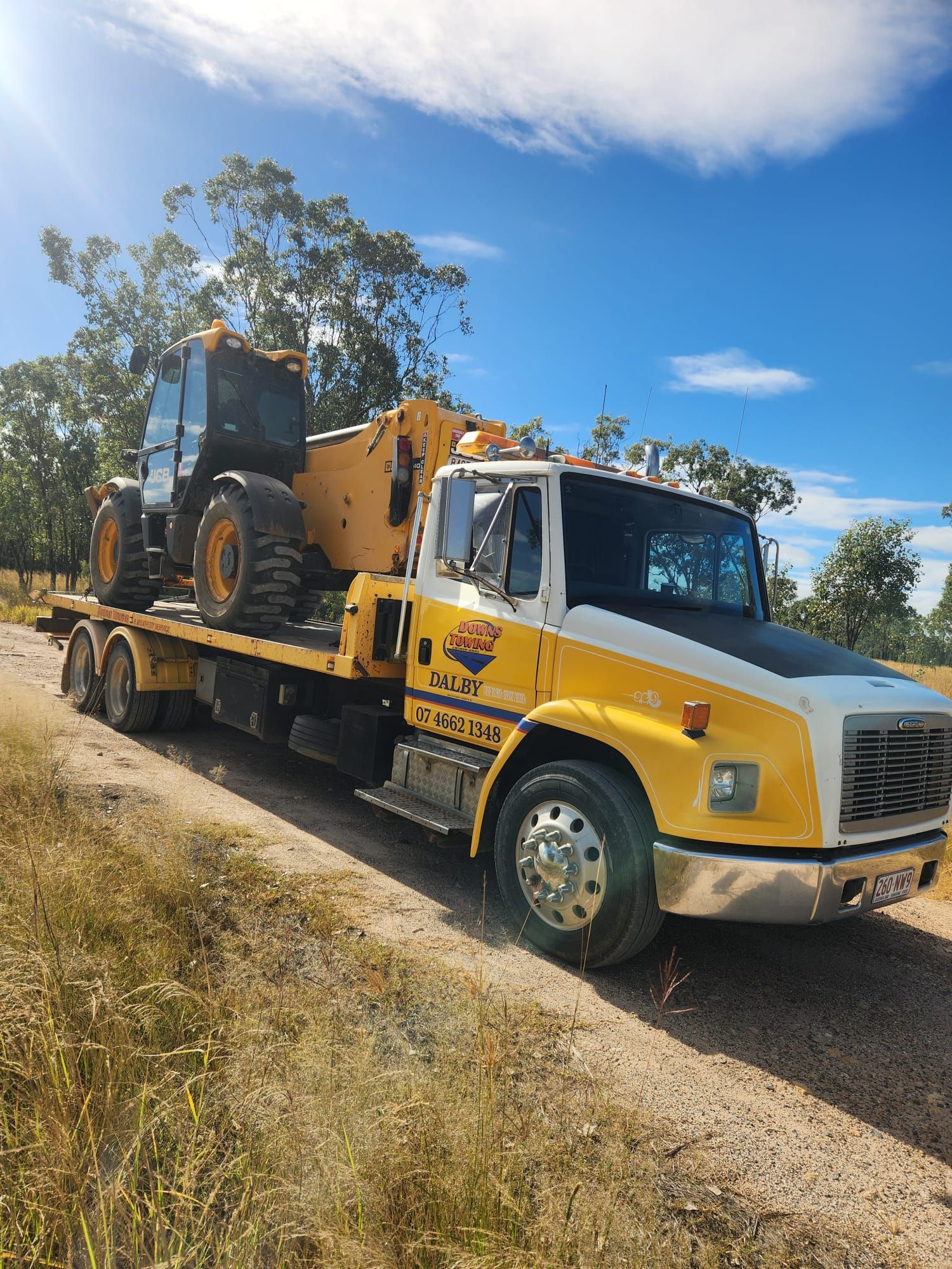 Yellow Heavy Truck Hauling A Tractor On A Flatbed Trailer Along A Rural Road Under A Blue Sky — Downs Towing & Recovery Service in Dalby, QLD