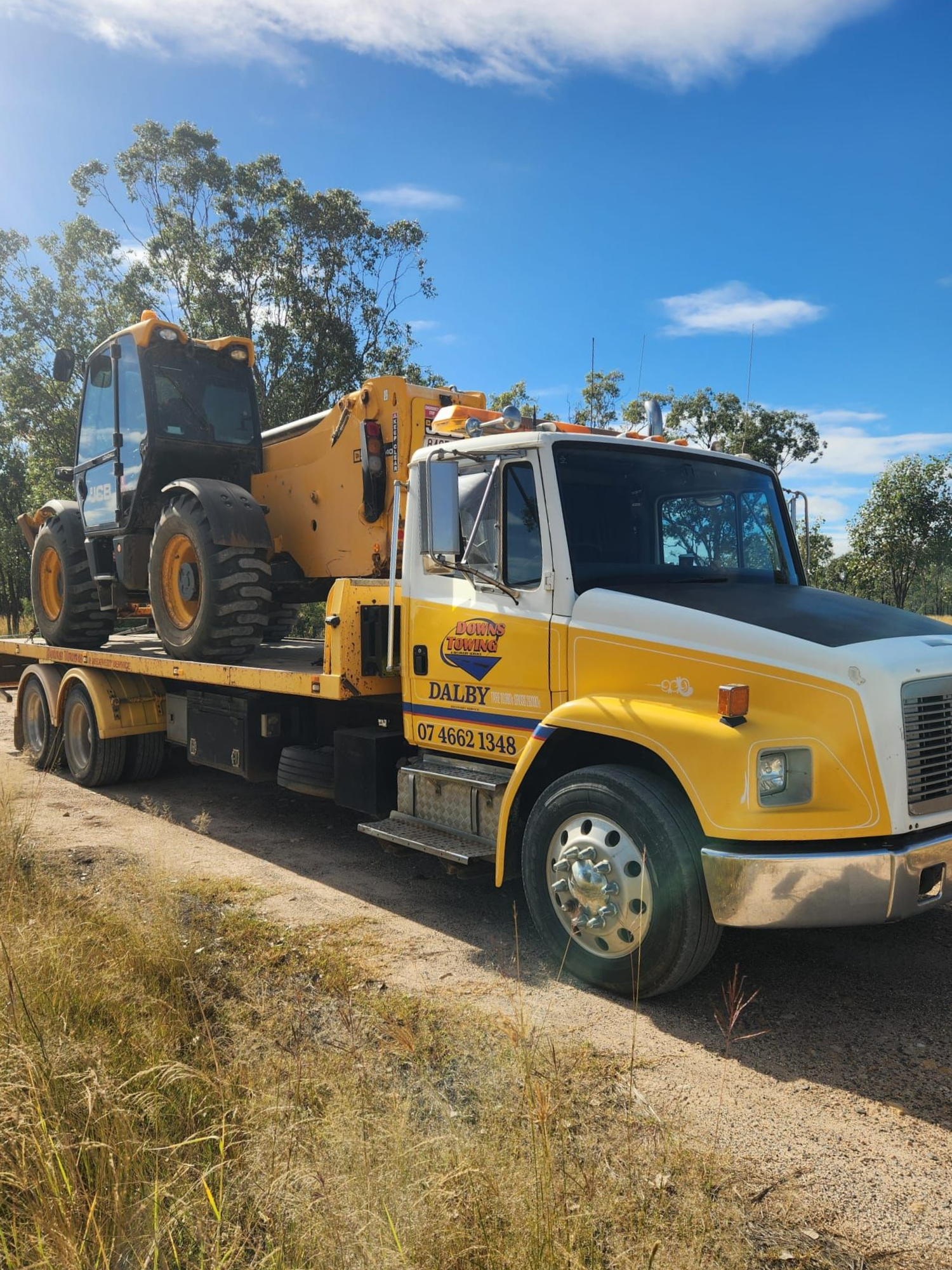 Yellow Tow Truck Carrying A Black Skid Steer On A Rural Dirt Road Under A Blue Sky — Downs Towing & Recovery Service in Dalby, QLD