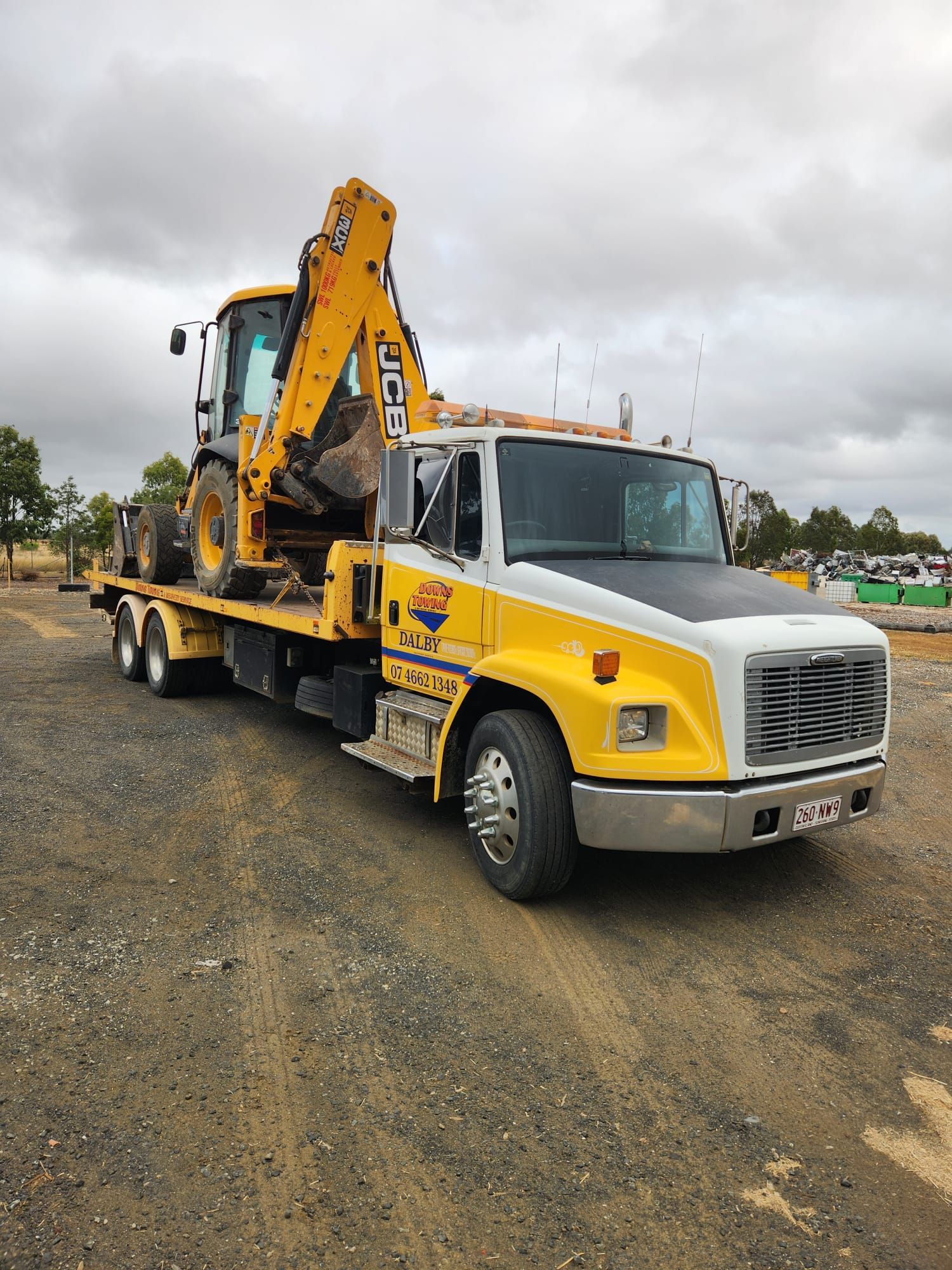 Yellow flatbed tow truck carrying a backhoe on a gravel lot under cloudy skies — Downs Towing & Recovery Service in Miles, QLD