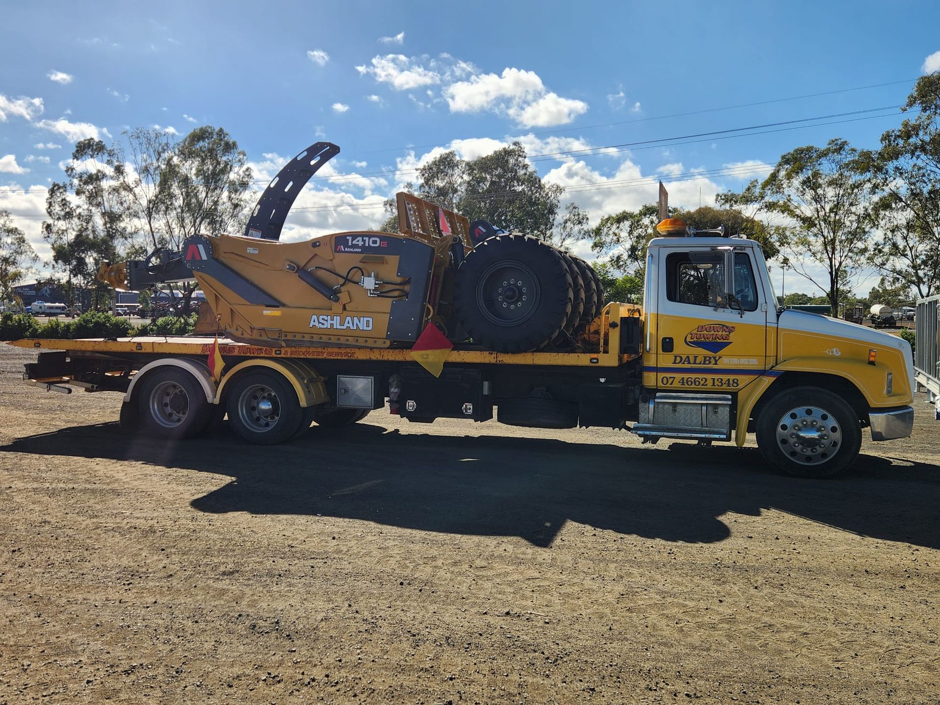 Yellow Tow Truck Carrying A Skid-steer Loader On A Trailer, Parked On A Gravel Lot Under A Blue Sky — Downs Towing & Recovery Service in Chinchilla, QLD