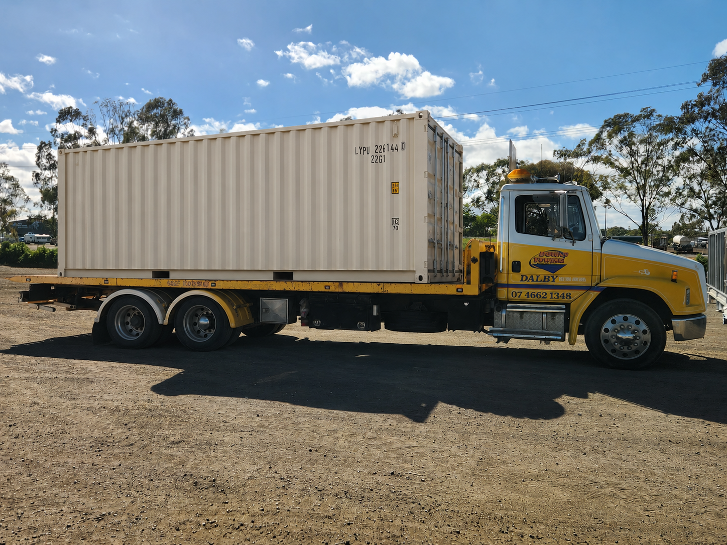 Yellow Flatbed Truck Carrying A Large Beige Shipping Container On A Gravel Lot — Downs Towing & Recovery Service in Dalby, QLD