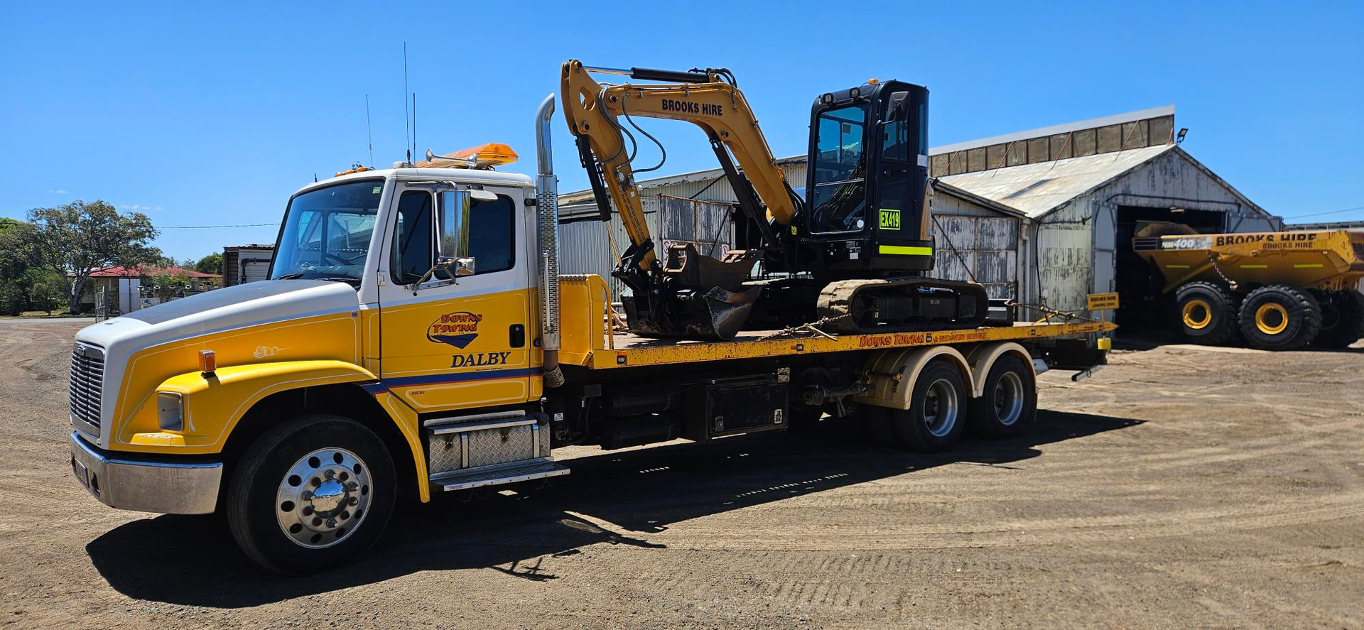 Yellow And White Semi-truck With A Green Machine On A Flatbed Trailer In A Sunny Setting — Downs Towing & Recovery Service in Dalby, QLD