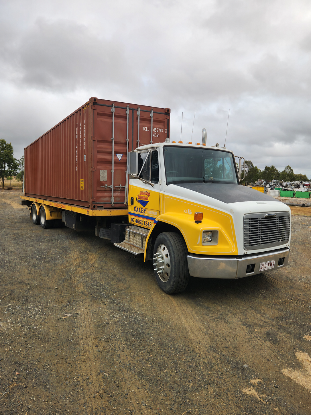 Yellow Semi-truck Hauling A Red Shipping Container On A Gravel Lot Under Cloudy Skies — Downs Towing & Recovery Service in Dalby, QLD