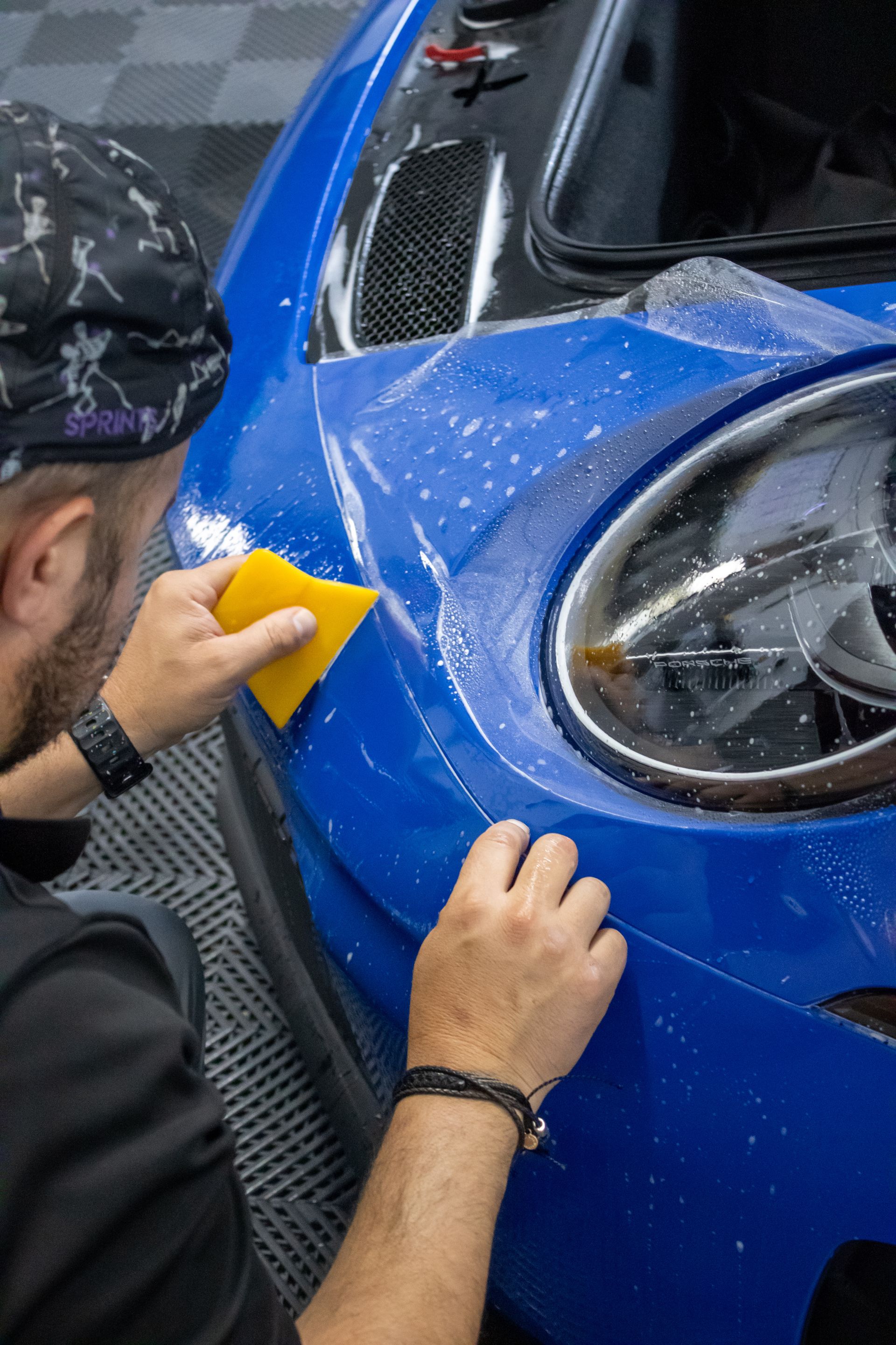 Person applying a yellow cloth to a blue car’s front fender near the headlight
