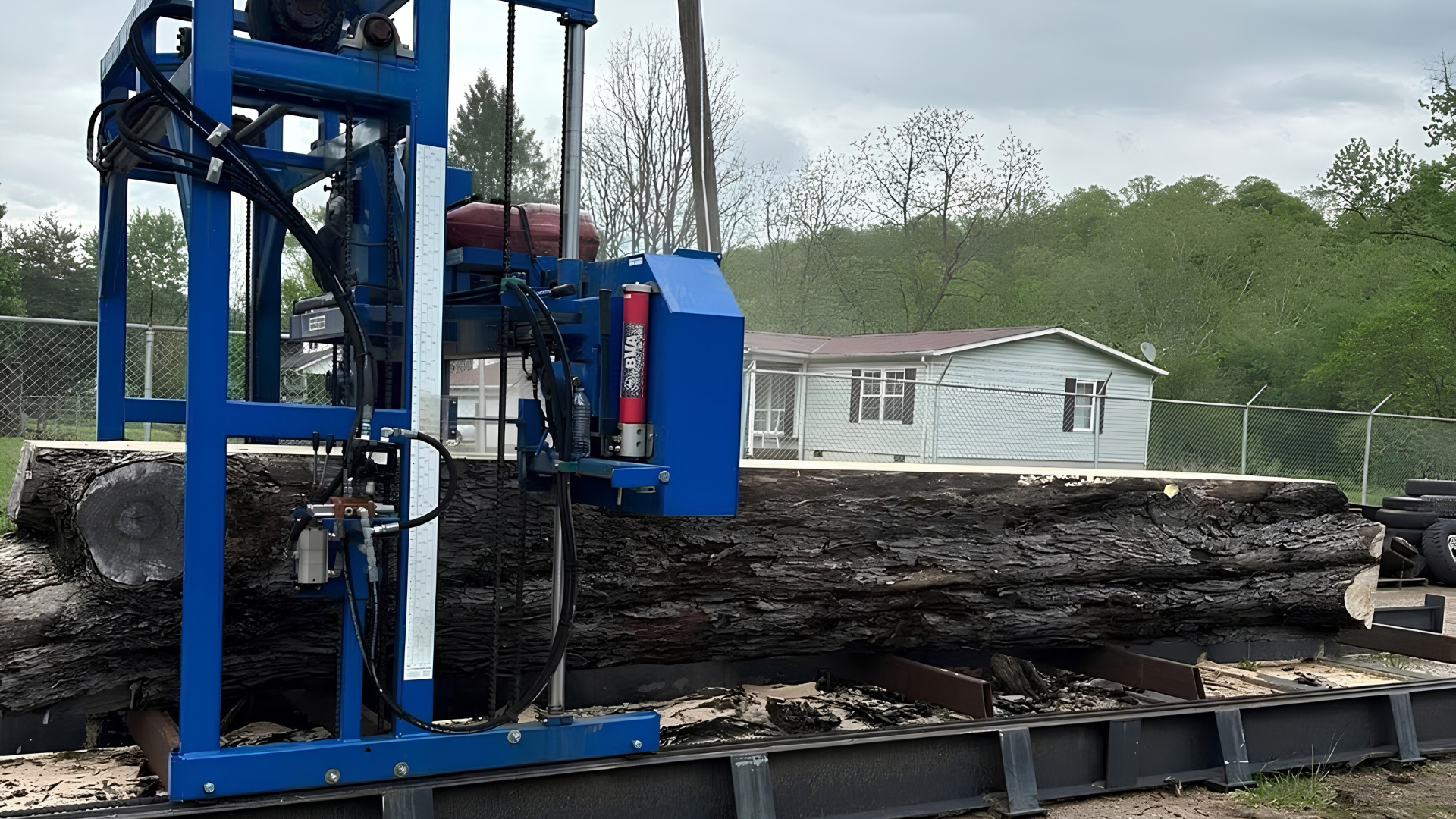 Blue log-splitting machine loading a large fallen log outdoors near a fence and shed, work of Specialty Slabwood Creations