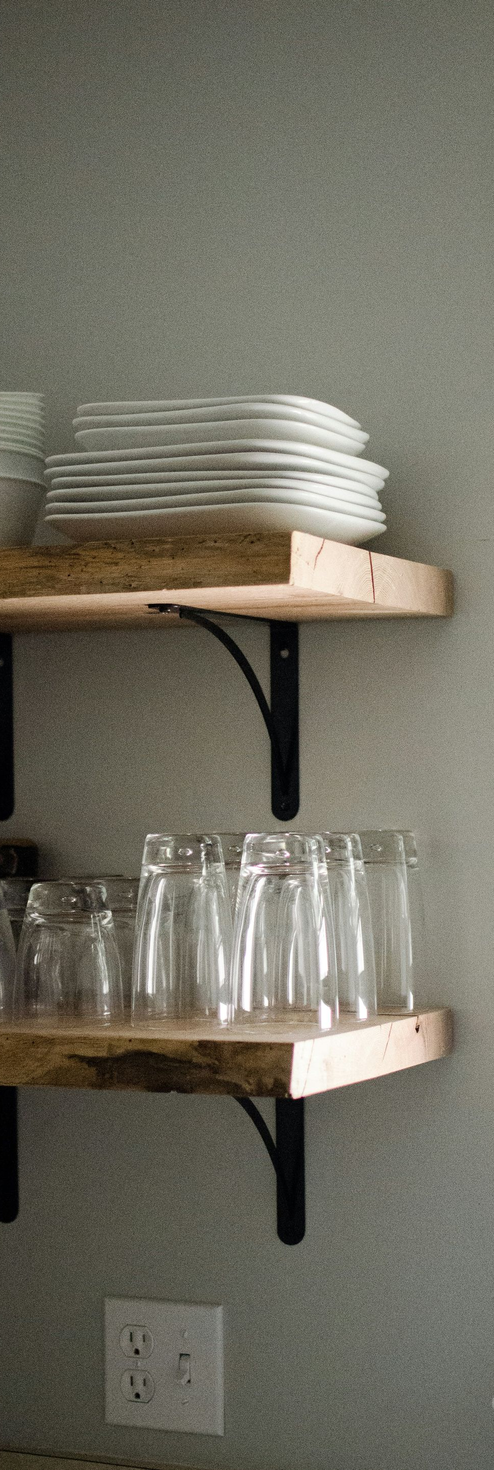 Kitchen shelves with stacked white plates on top and drinking glasses below, against a light wall.