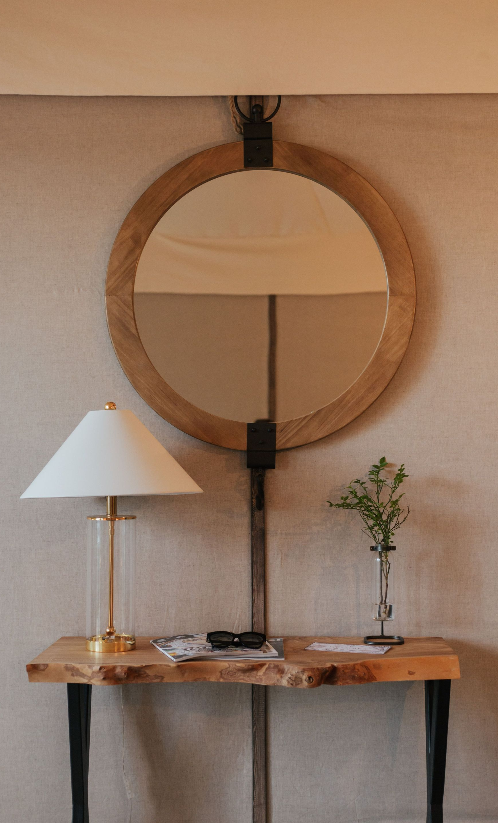 Round wood mirror above rustic console with white lamp and small plant on a beige wall