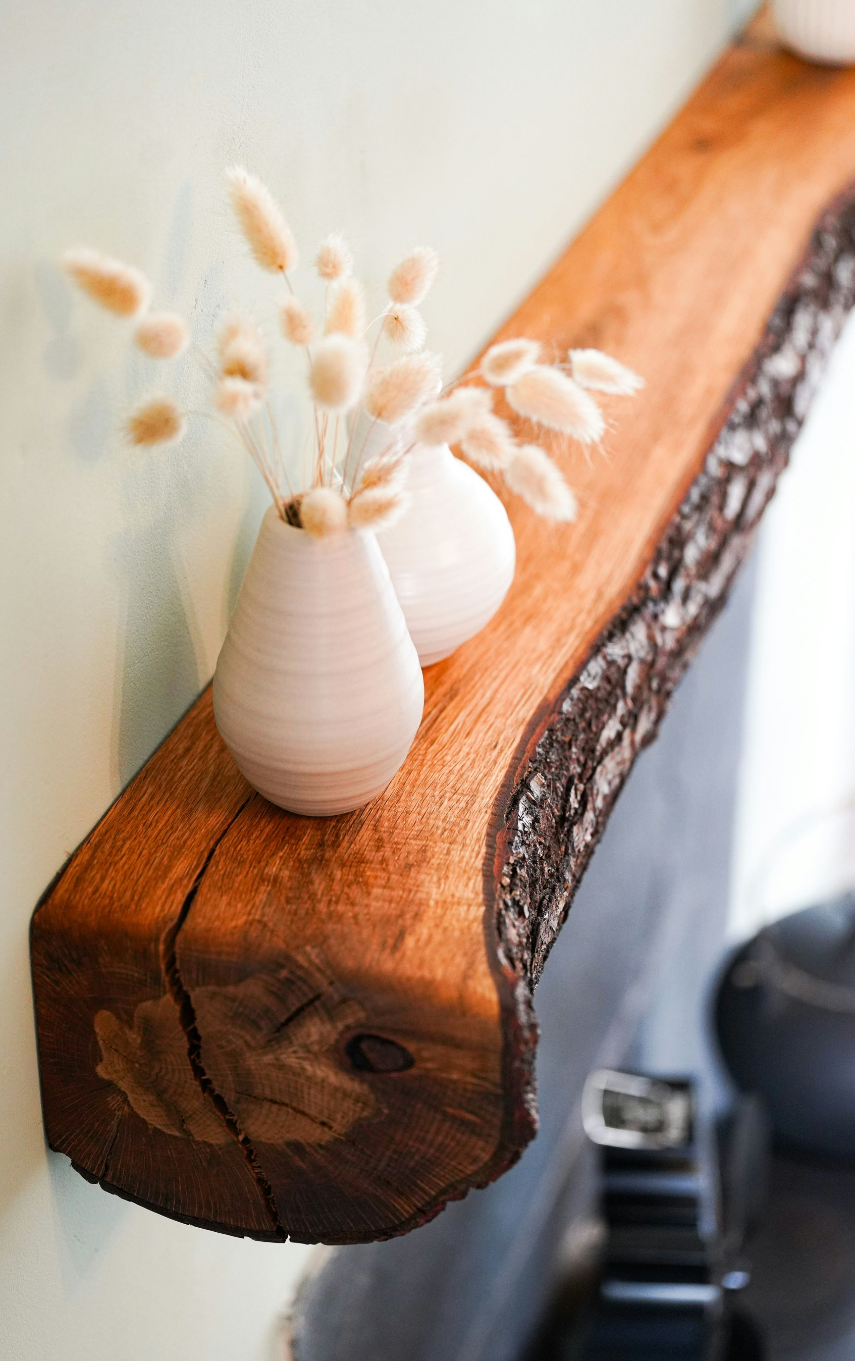 Small white vase with dried stems on a rustic wooden shelf against a light wall.