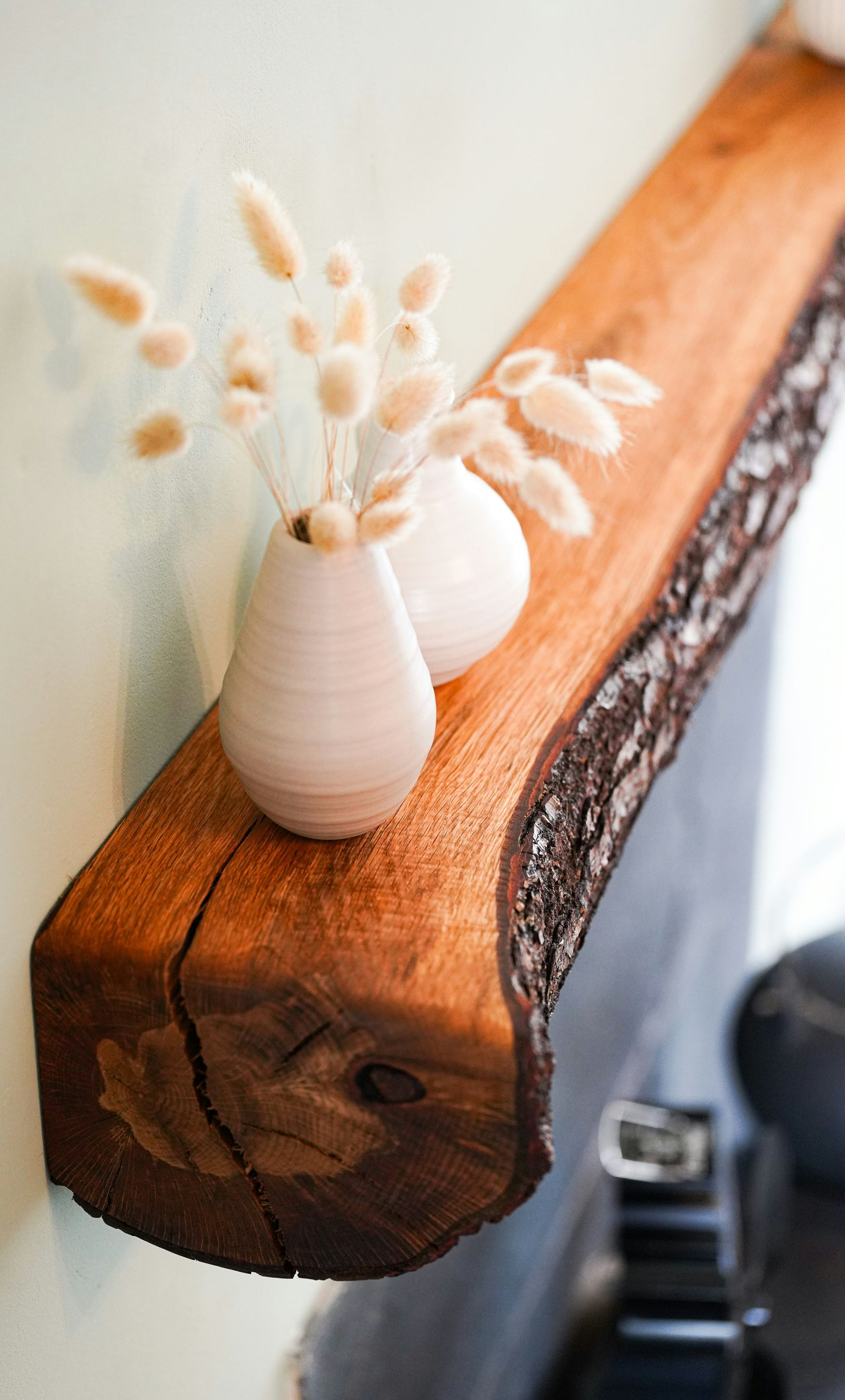 Small white vase with dried stems on a rustic wooden shelf against a light wall.