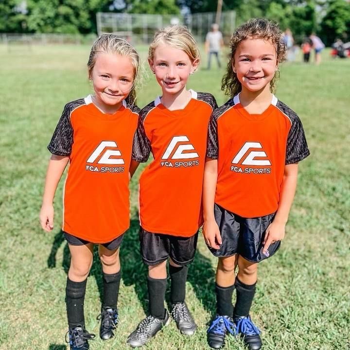 Three youth soccer players in orange uniforms posing on a grassy field