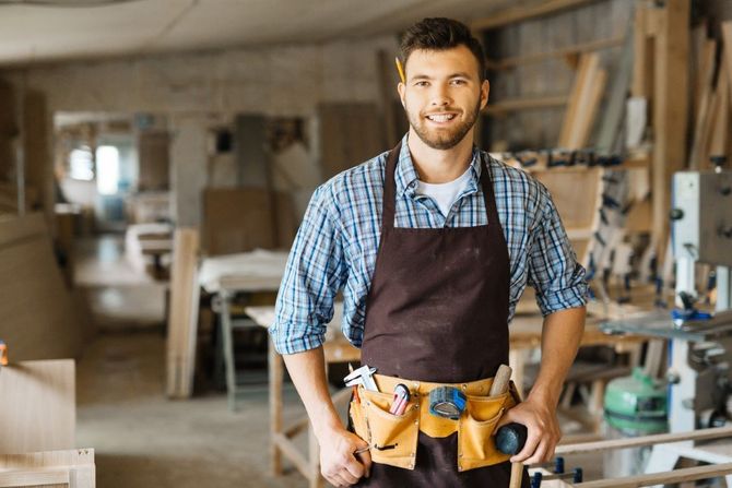 Man in a blue plaid shirt and brown apron standing in a woodshop with tools and machinery behind him