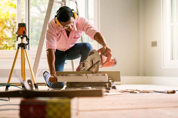 Person kneeling on a wooden floor, measuring and cutting a board with tools in a bright room.