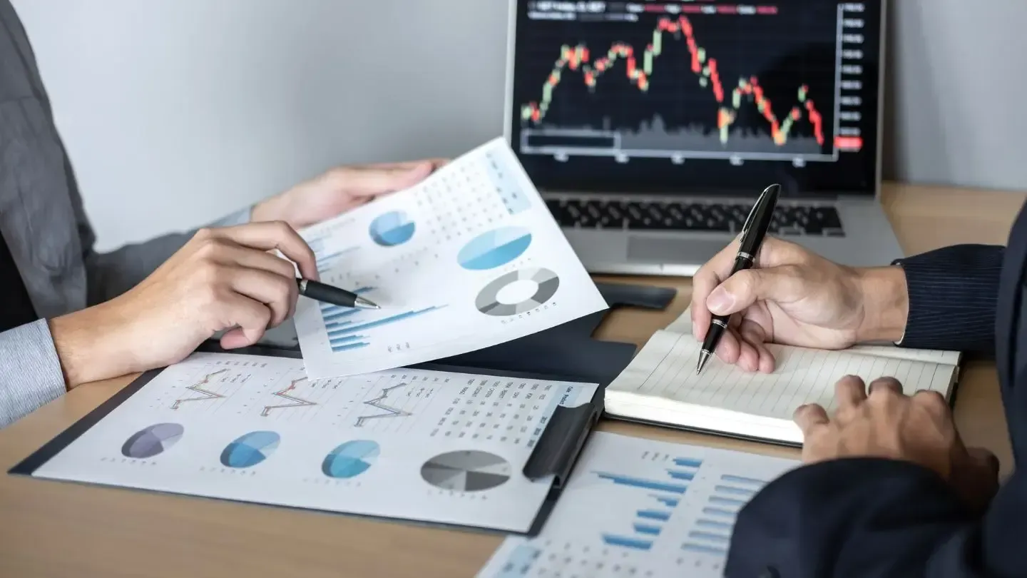 Two people reviewing financial charts and graphs at a desk with a laptop displaying stock trends.
