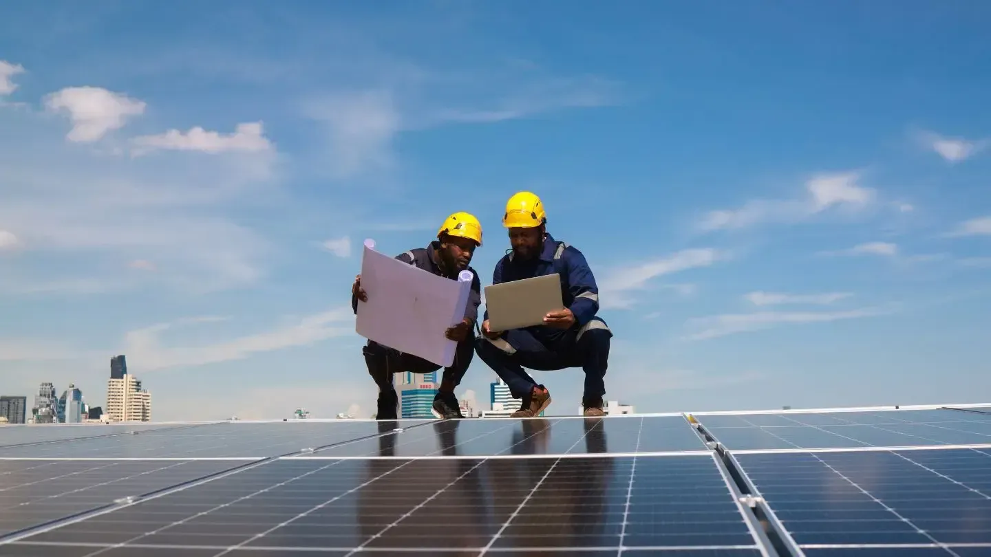 Two workers in hard hats examine a blueprint and laptop on a solar panel array under a blue sky.