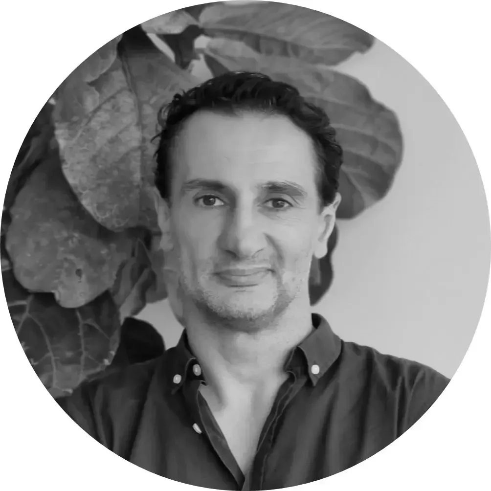 A man with dark hair and a subtle smile, wearing a button-down shirt, poses in front of a leafy plant.