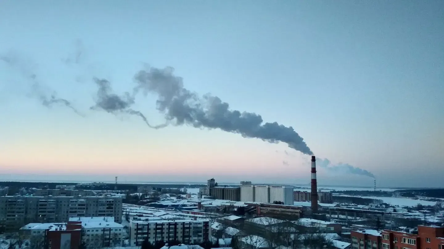 Smoke billows from a tall industrial chimney over a snow-covered cityscape at dusk.