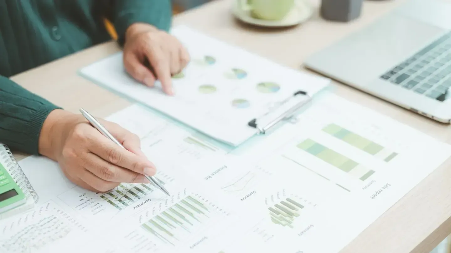 Person analyzing financial reports with pen, charts, and laptop on a desk.