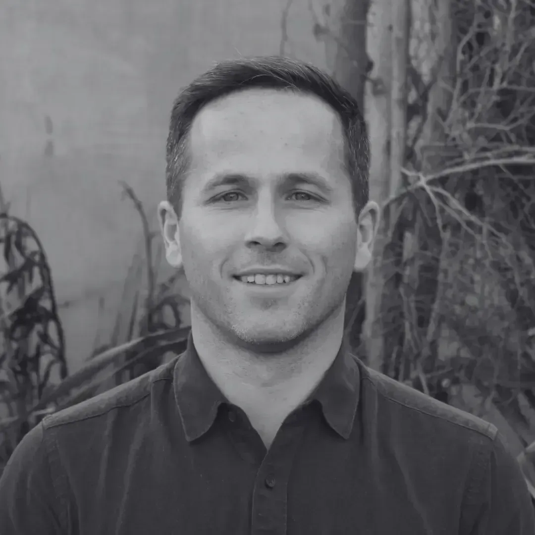 Man with short dark hair smiles at the camera, wearing a dark button-down shirt. Background is outdoors with trees.