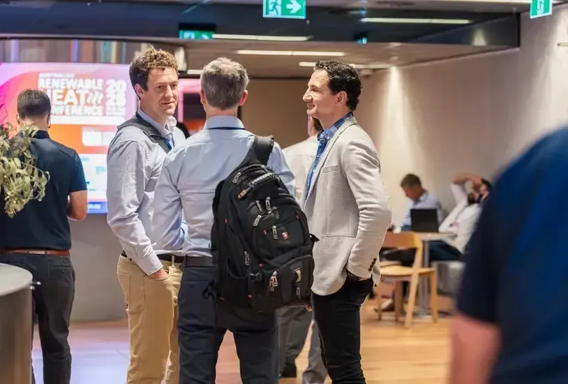 Three men in business attire conversing at a conference, smiling, in a well-lit hallway setting.