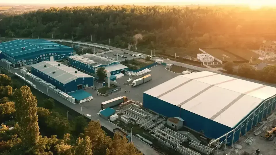 Aerial view of an industrial complex with blue-roofed buildings, trucks, and a parking lot, near a forest.