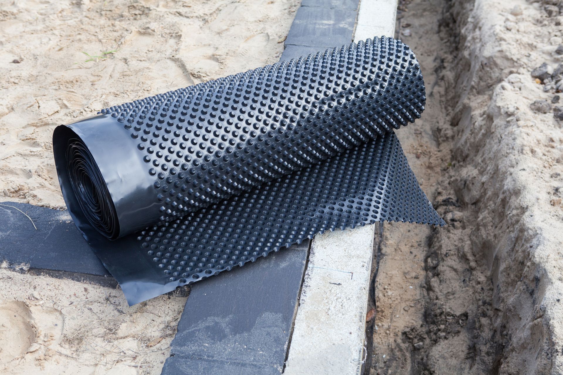 A roll of drainage mat is laying on the ground next to a concrete curb.