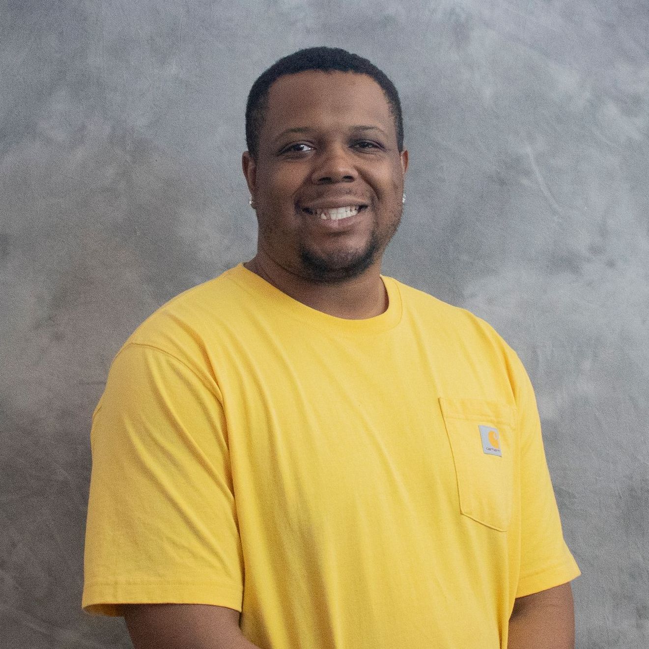 A man in a yellow shirt is standing in front of a grey wall.