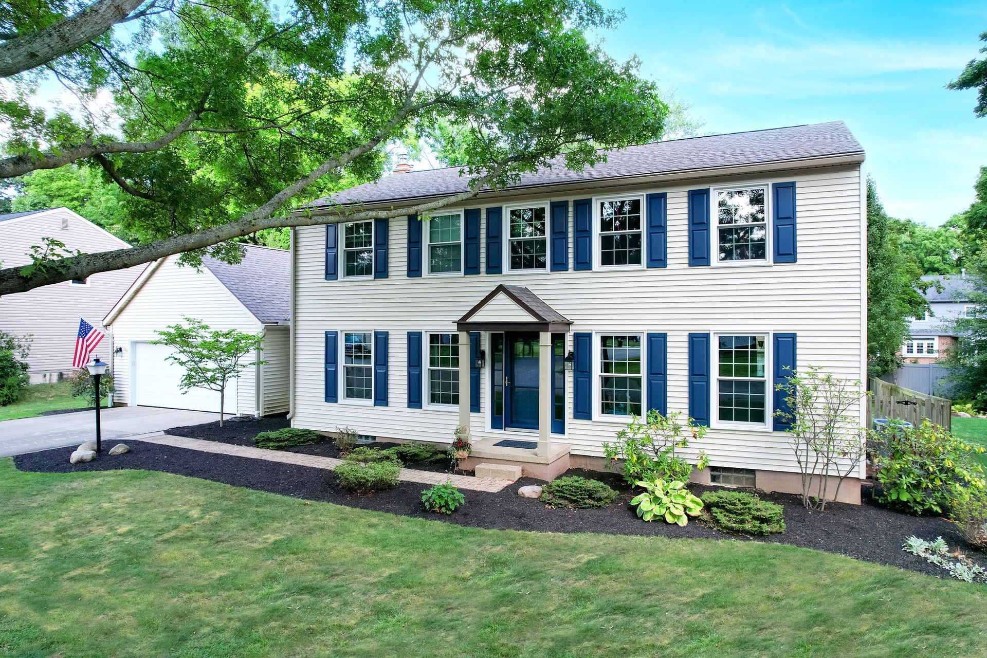 Two-story white house with blue shutters, front lawn, and American flag on a sunny day