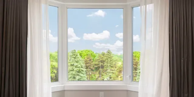 Bright bay window with sheer curtains overlooking green trees and a blue sky