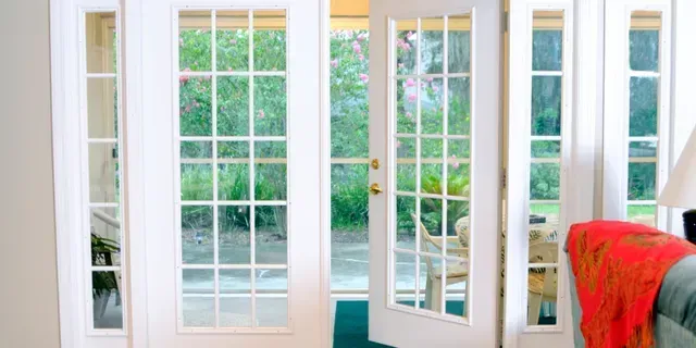 White French doors with glass panes open to a green garden, with a red blanket on a chair inside.