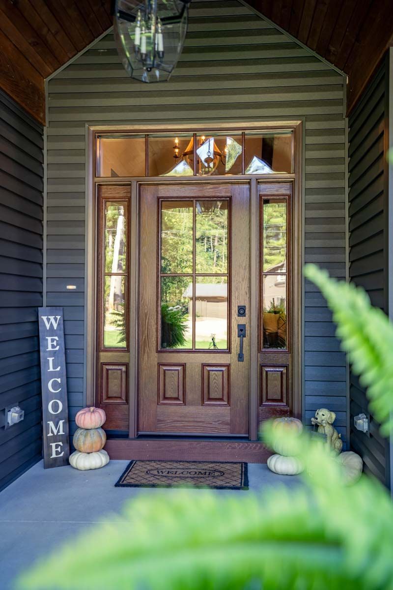 Front porch with open front door, welcome sign, and pumpkins, framed by dark siding and greenery.