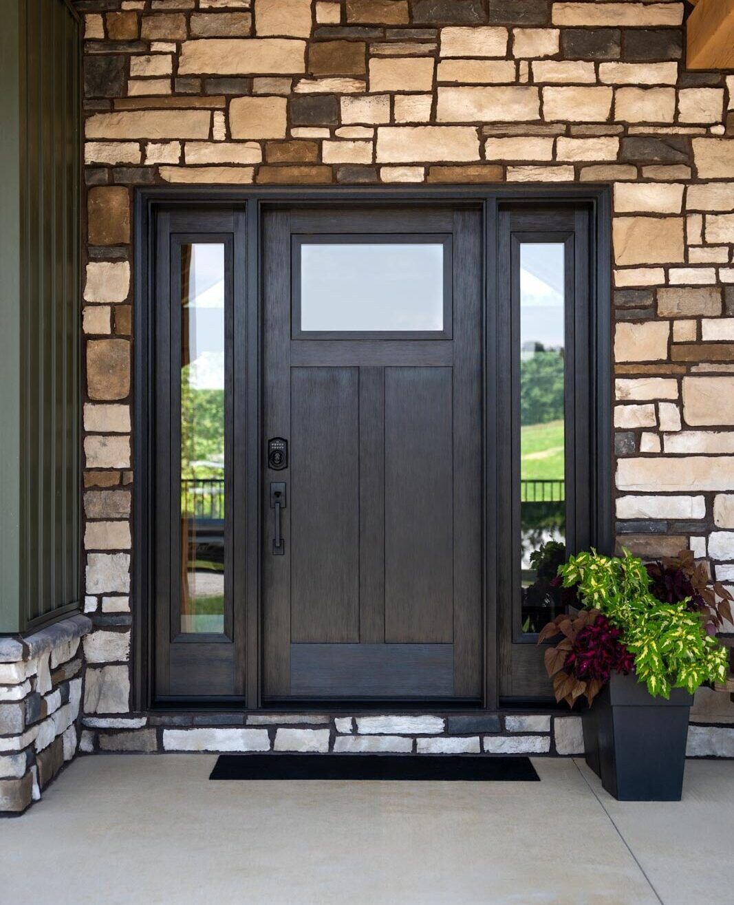 Stone-front entryway with a dark wood front door, sidelights, and potted plants on a porch