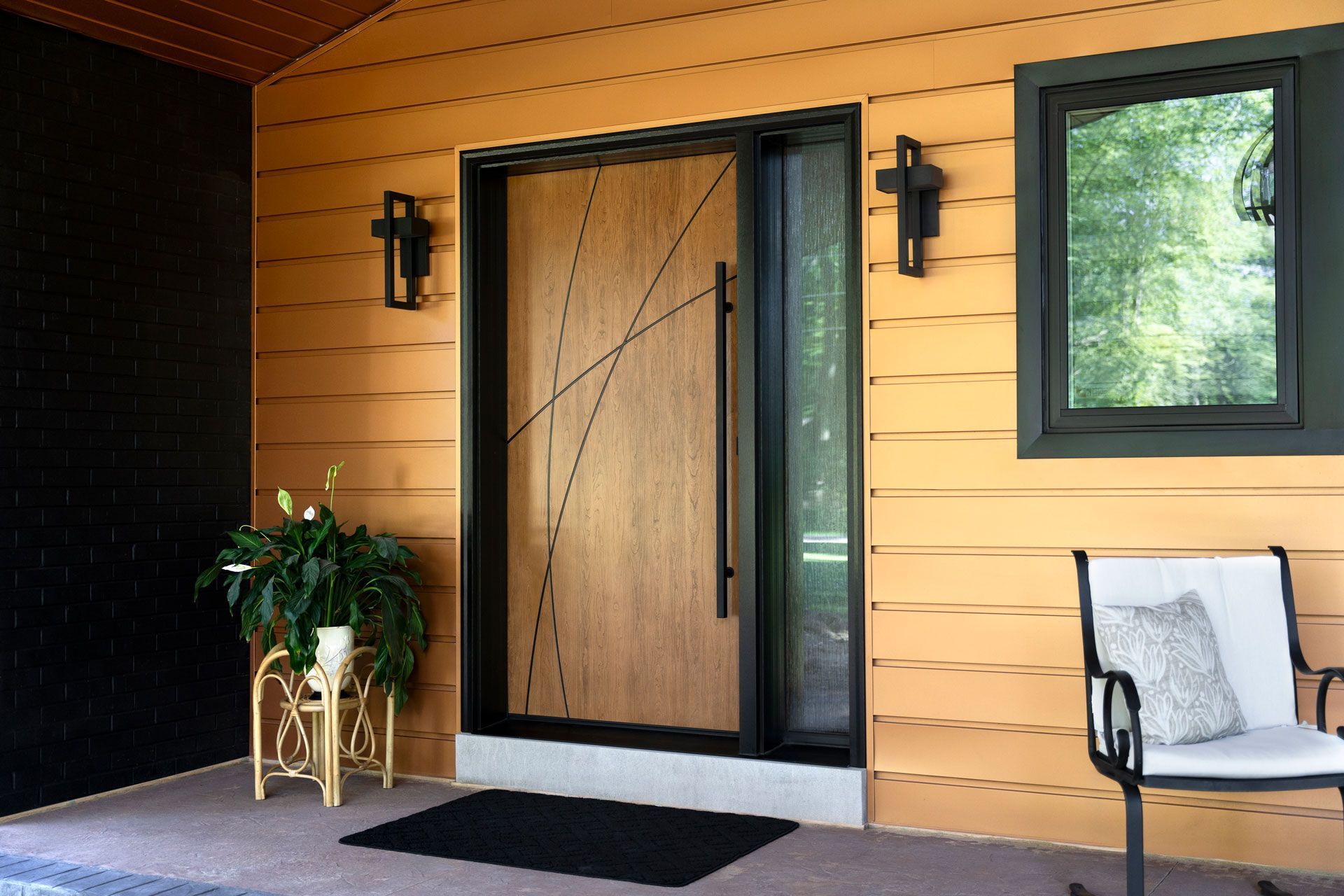 Covered front porch with wooden door, black trim, wall lanterns, potted plant, and chair on the right.